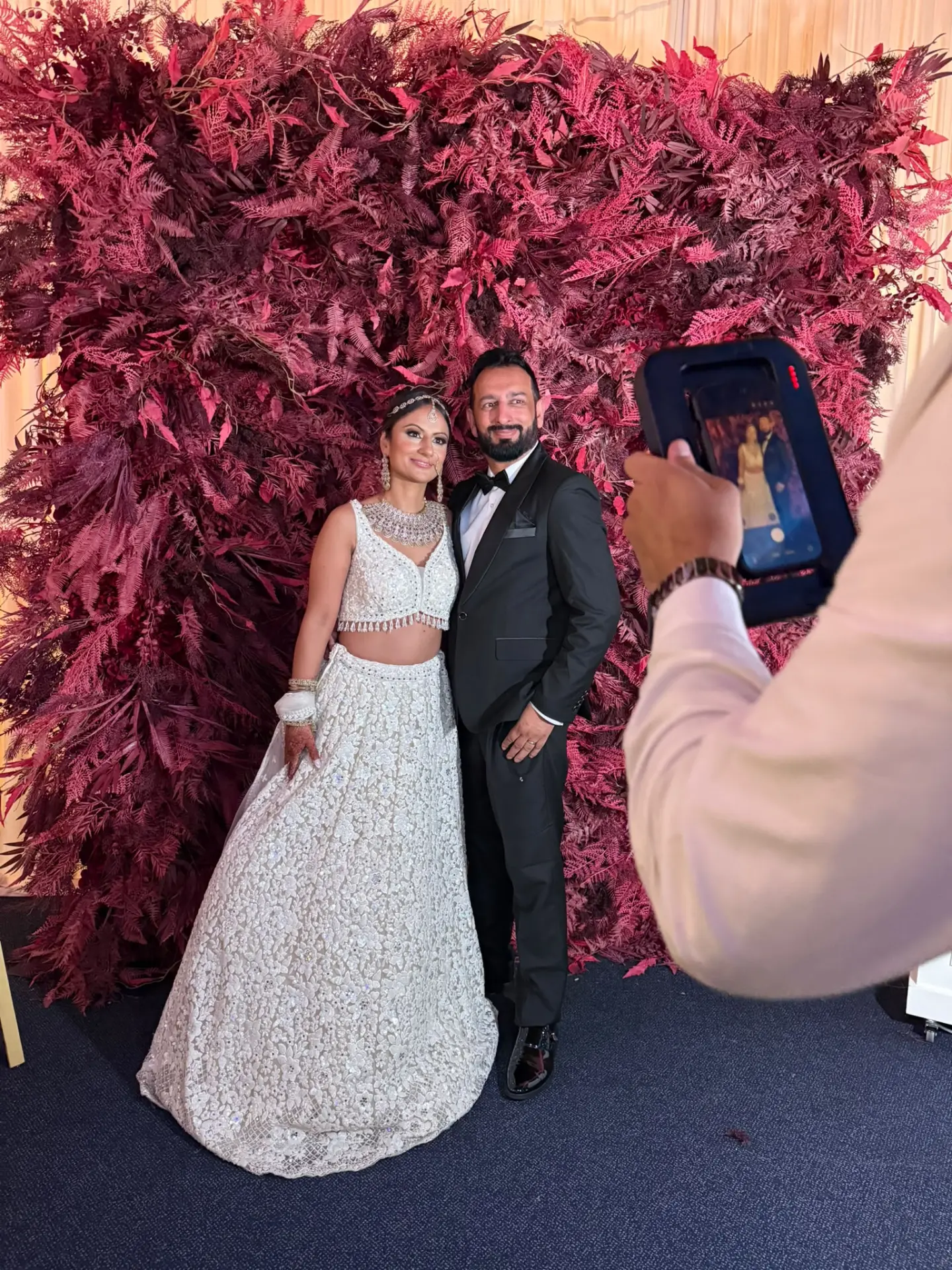 A couple being photographed in front of a red foliage wall at their reception