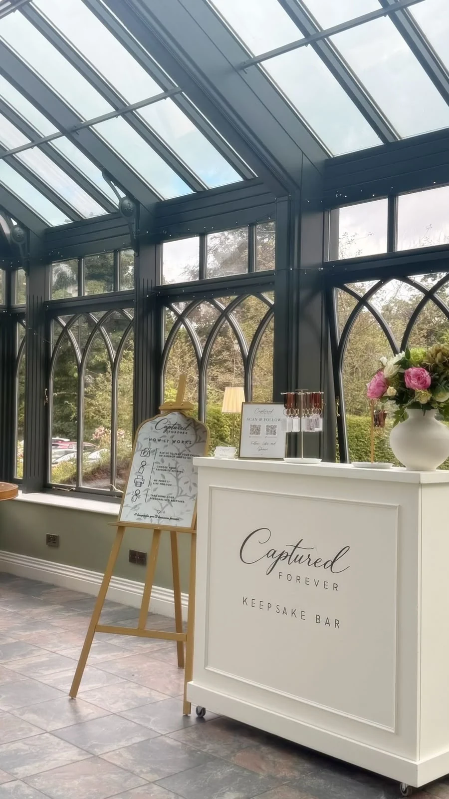 The Captured Forever Keepsake Bar with its How It Works easel set beneath the glass roof of a glasshouse venue