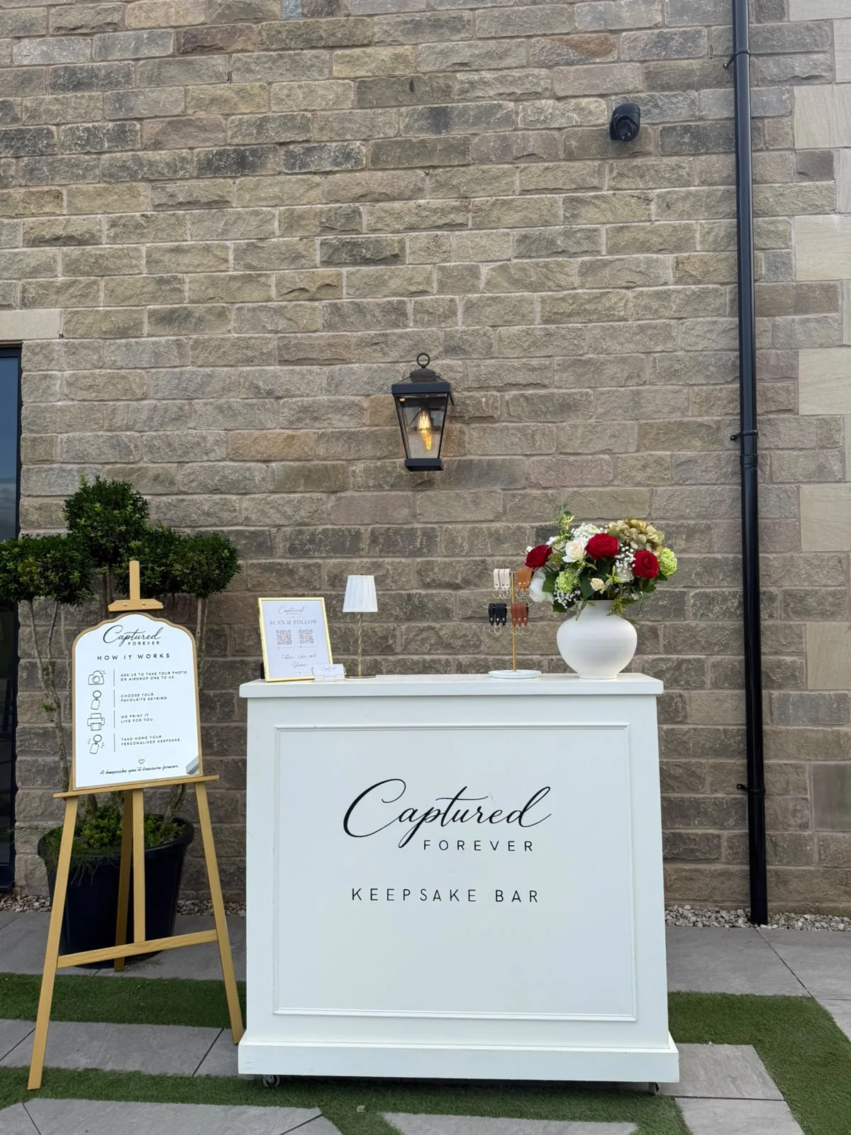 The Captured Forever Keepsake Bar and How It Works easel set against a brick venue wall with flowers and keyring displays